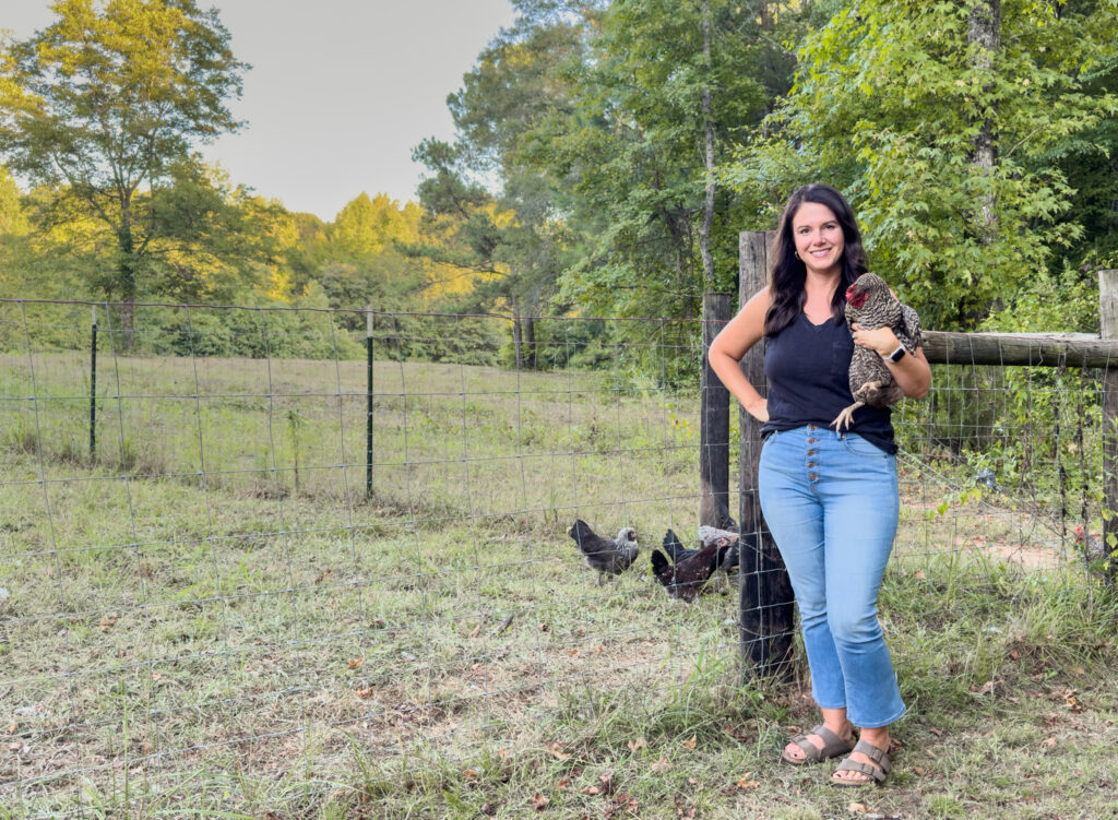 Woman with brown hair holding barred rock chicken in front of fence with other chickens around. 