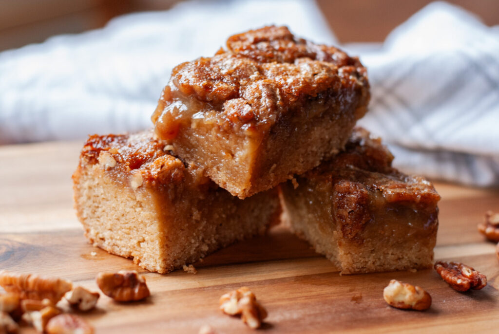 pean pie blondies squares stacked on top of each other surrounded by pecans. 